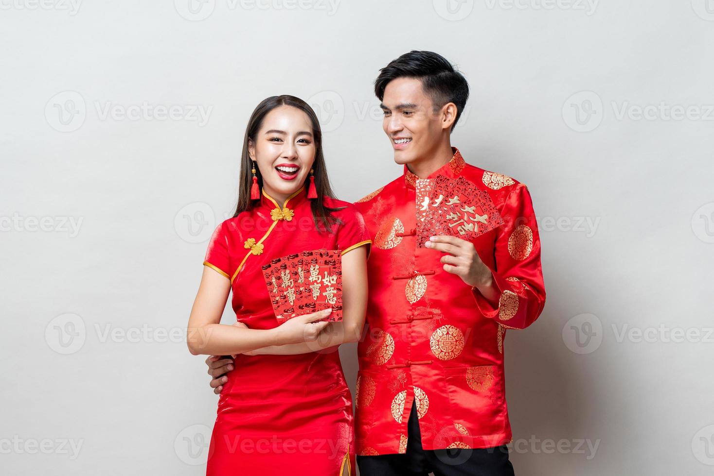 feliz pareja asiática con trajes orientales tradicionales sosteniendo sobres rojos o ang pao con fondo gris claro para conceptos de año nuevo chino, los textos significan gran suerte, gran beneficio foto