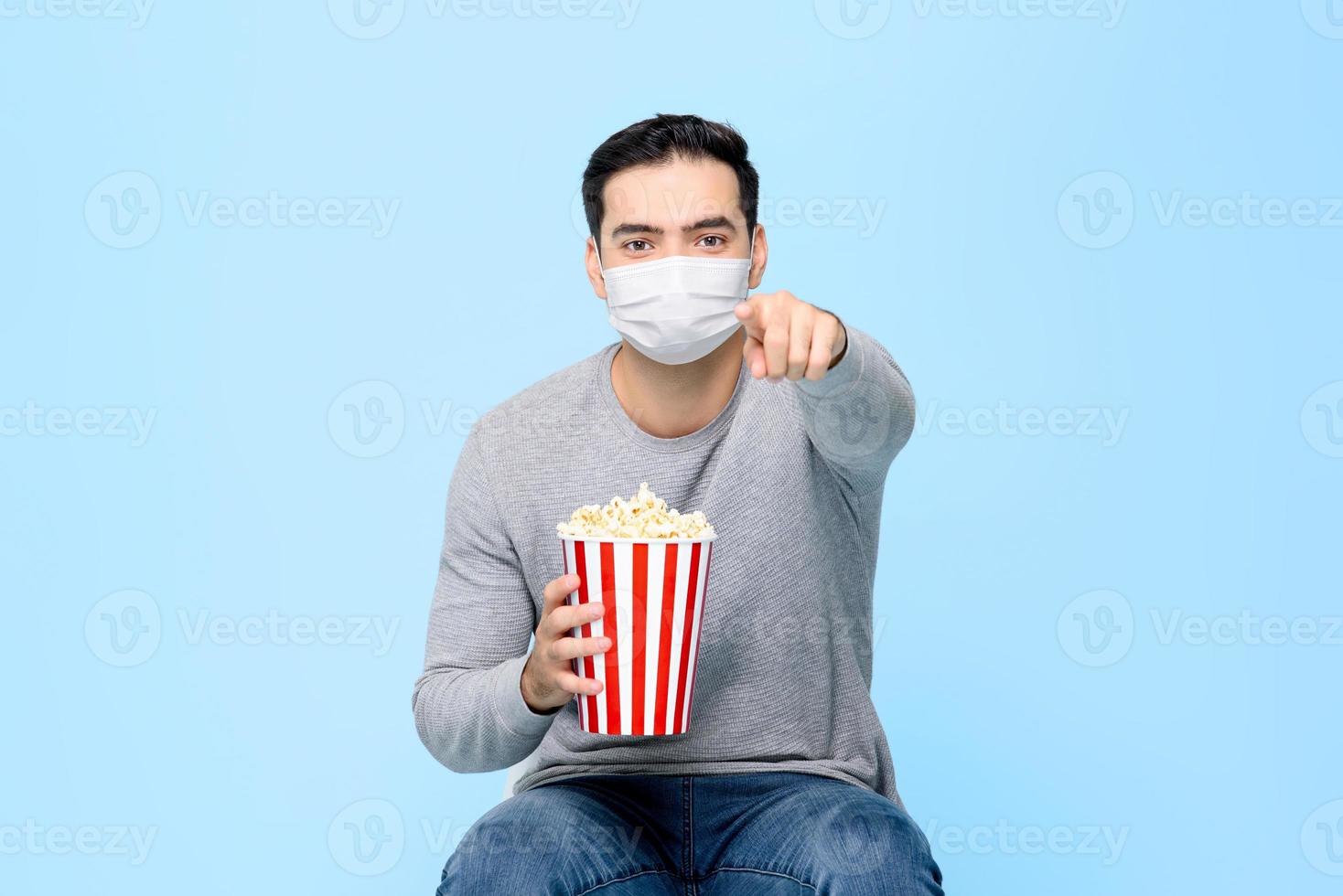 un joven con palomitas de maíz se protege usando una mascarilla médica mientras disfruta viendo una película aislada en un fondo azul claro foto