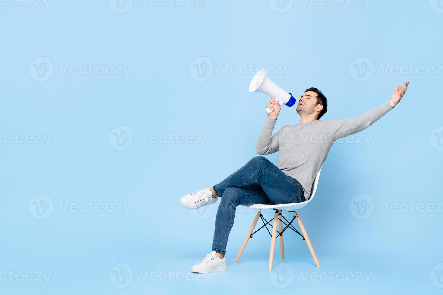 Portrait of young handsome Caucasian man sitting and cheerfully announcing on megaphone with one arm raised in isolated studio blue background photo