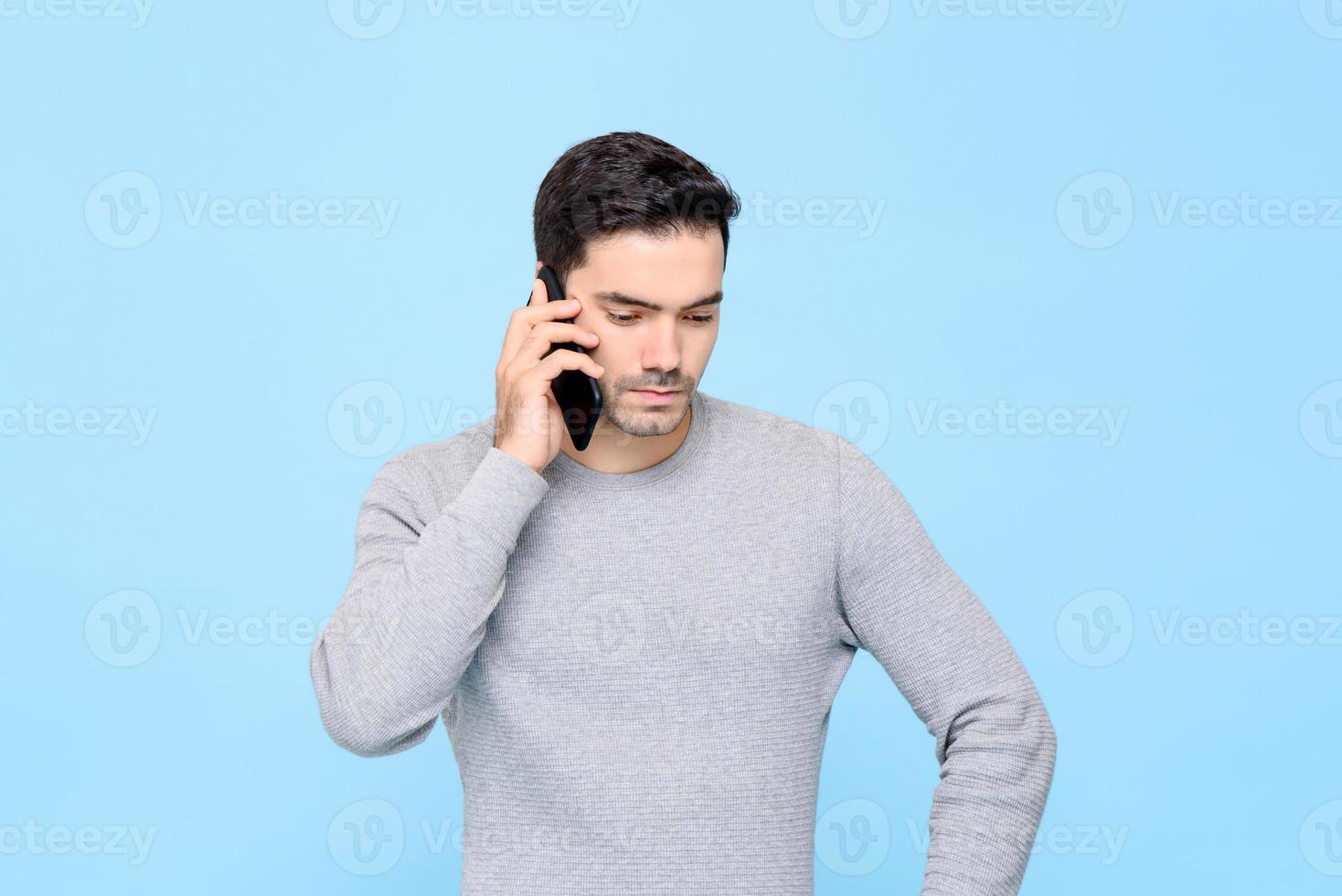 Portrait of serious and upset handsome Caucasian man calling on cellphone with unhappy facial expression in isolated studio blue background photo