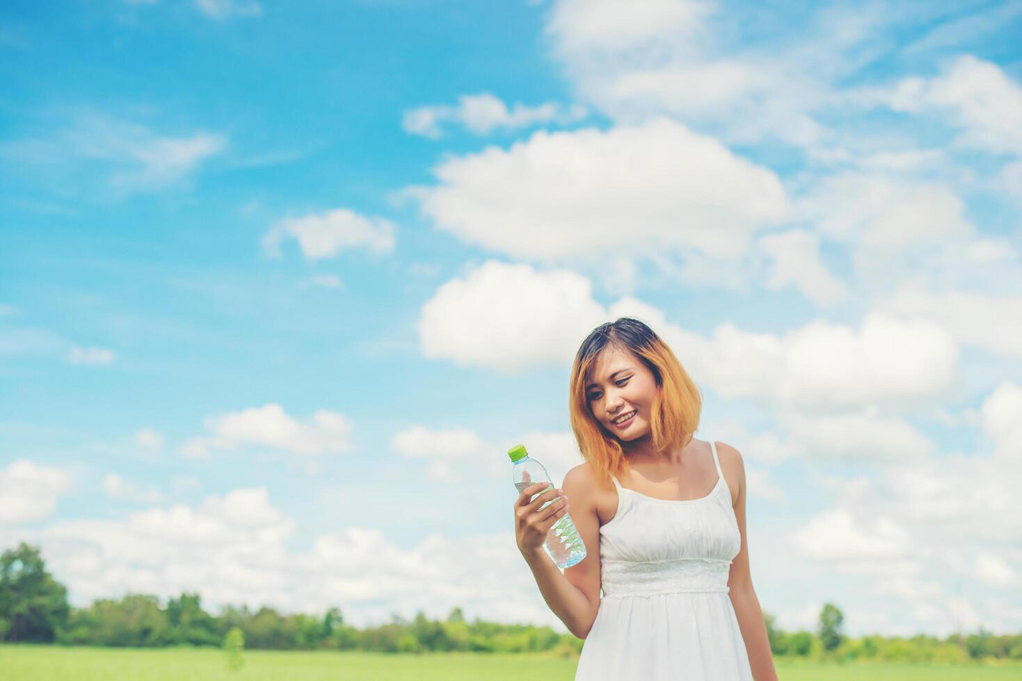 concepto de estilo de vida de las mujeres mujer joven y bonita con vestido blanco sosteniendo una botella de agua en los pastizales sonriente a la cámara se ve tan fresca, disfruta y feliz. foto