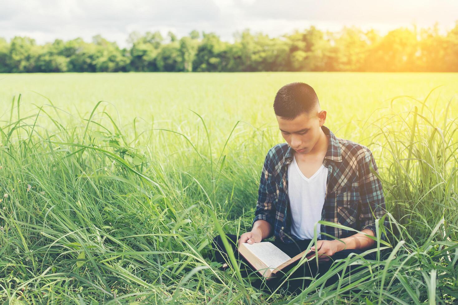 Young hipster man sitting on grassland reading book with nature around so happy. photo