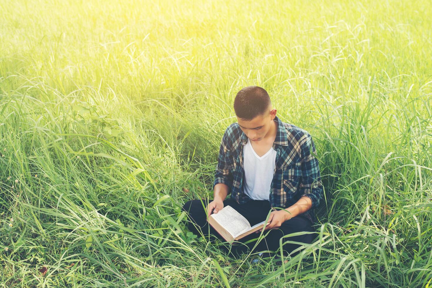 Young hipster man sitting on grassland reading book with nature around so happy. photo
