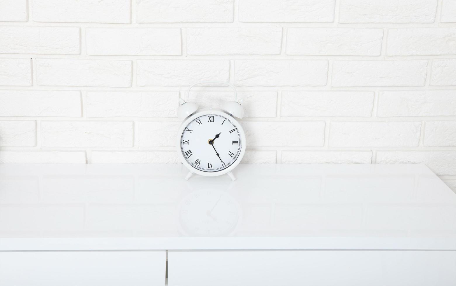 Retro alarm clock with twenty five minutes past one o'clock near brick wall on white workplace. Selective focus photo