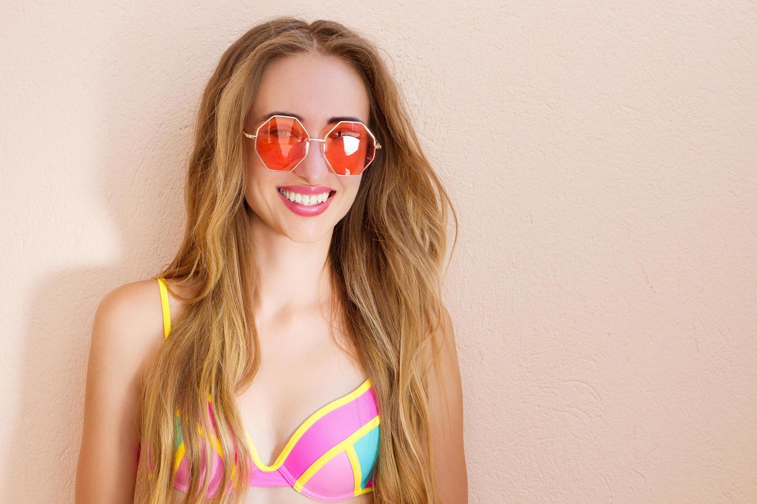 cerca de una chica feliz con gafas de sol rosas aisladas. vacaciones de verano y fin de semana divertido. concepto de verano. mujer joven sonriente en traje de baño de moda. enfoque selectivo. traje de playa de verano. copie el espacio foto