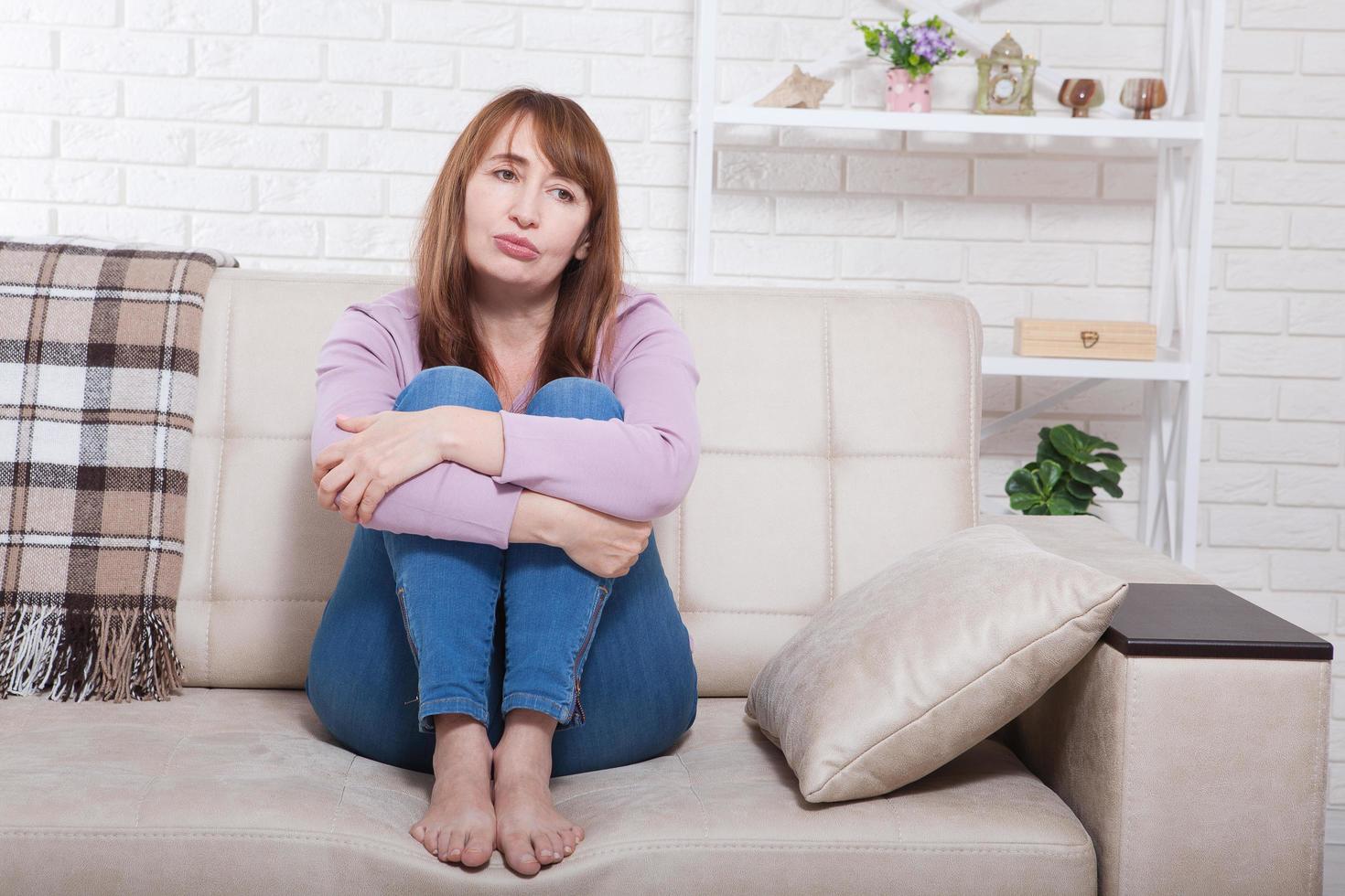 Depressed and sad middle aged woman sitting with clamped knees on bed, coach, sofa at home. Copy space and mock up. photo
