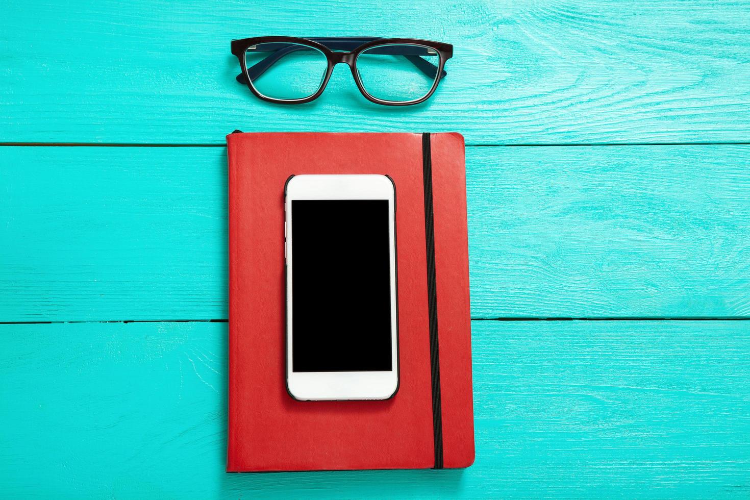Workplace with smartphone, coffee, glasses and red stylish notebook on blue wooden background. Top view photo