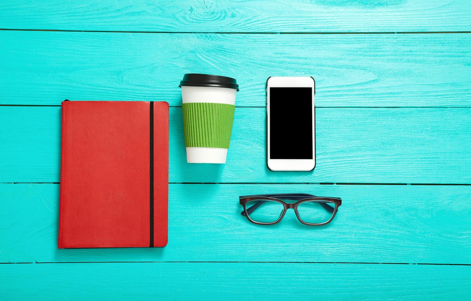 Workplace with smartphone, coffee, glasses and red stylish notebook on blue wooden background. Top view photo