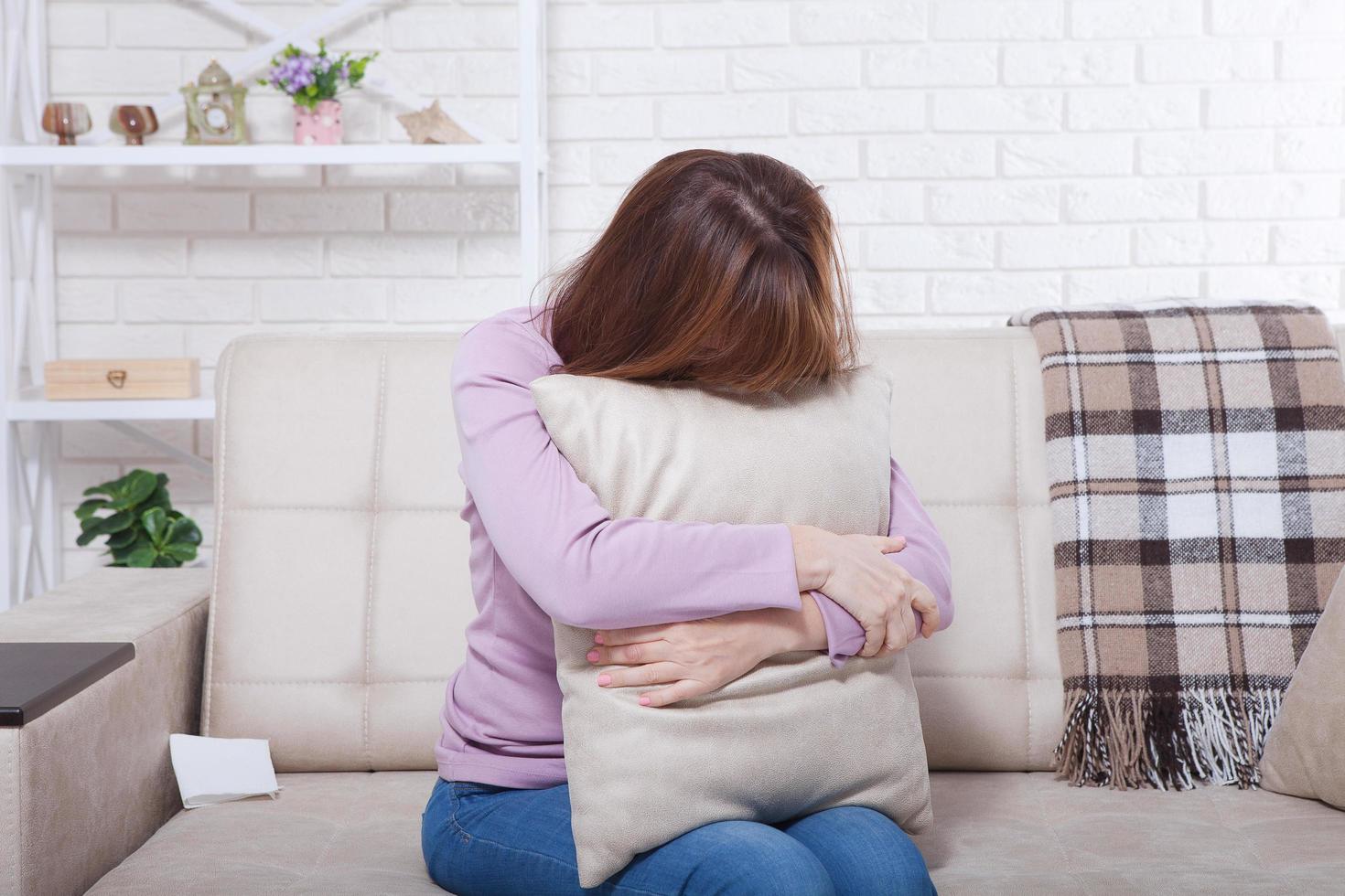 hermosa mujer morena de mediana edad sosteniendo una almohada y llorando en el sofá. fondo de casa, interior. tiempo de la menopausia. copie el espacio foto