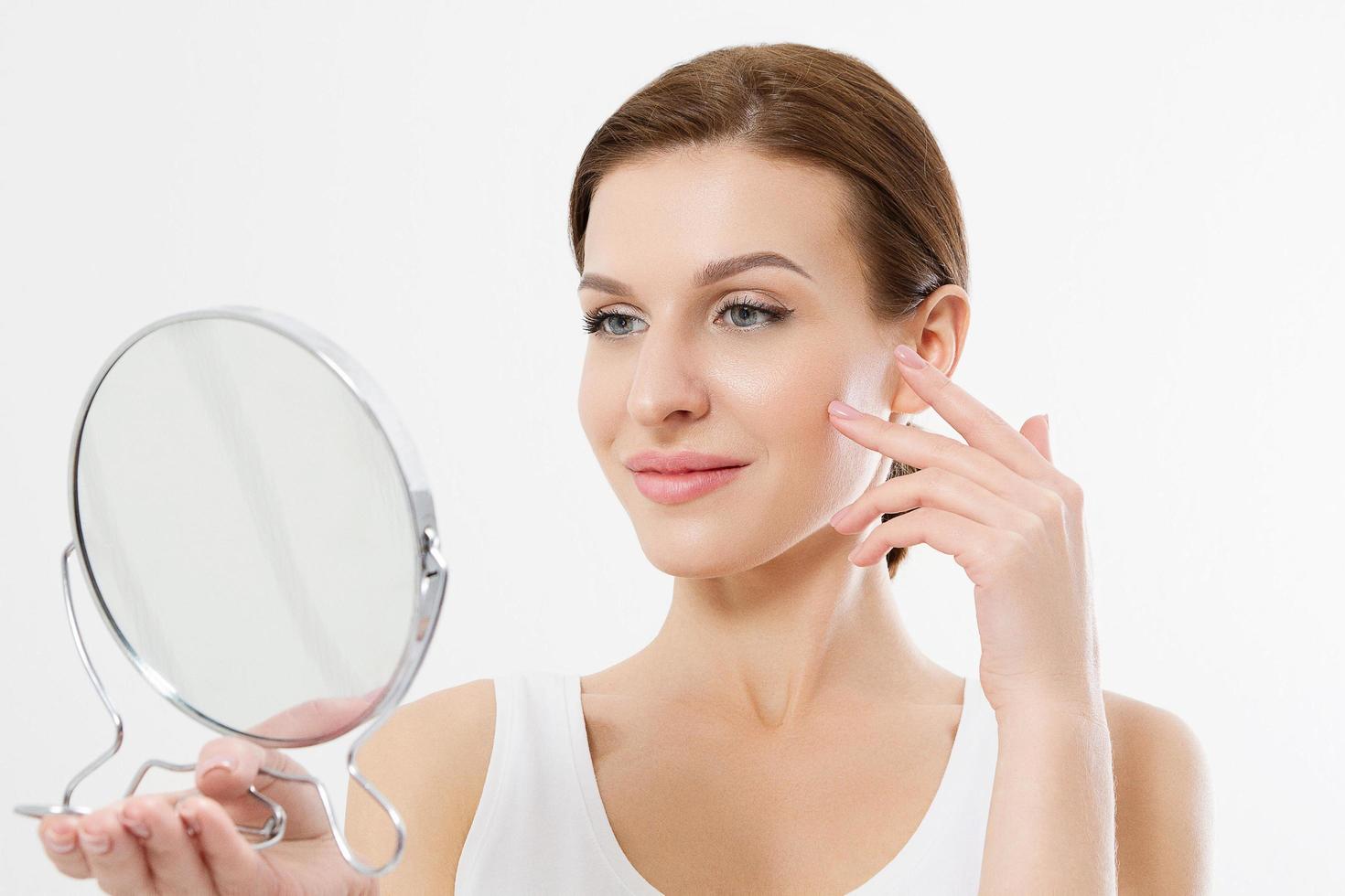 Smiling young woman with white teeth looking in mirror isolated on white background. Skin care and anti aging concept. Beauty and body health. Copy space photo