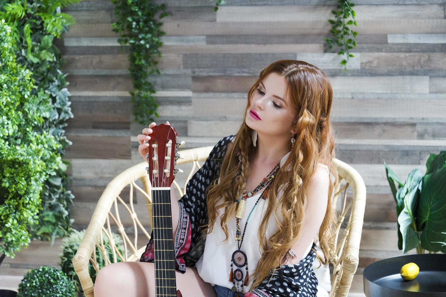 A beautiful girl with red curly hair sits on a chair with a seven-string guitar. photo