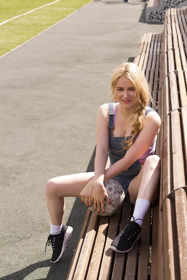 The girl sits on the bench at the football field with the ball. photo