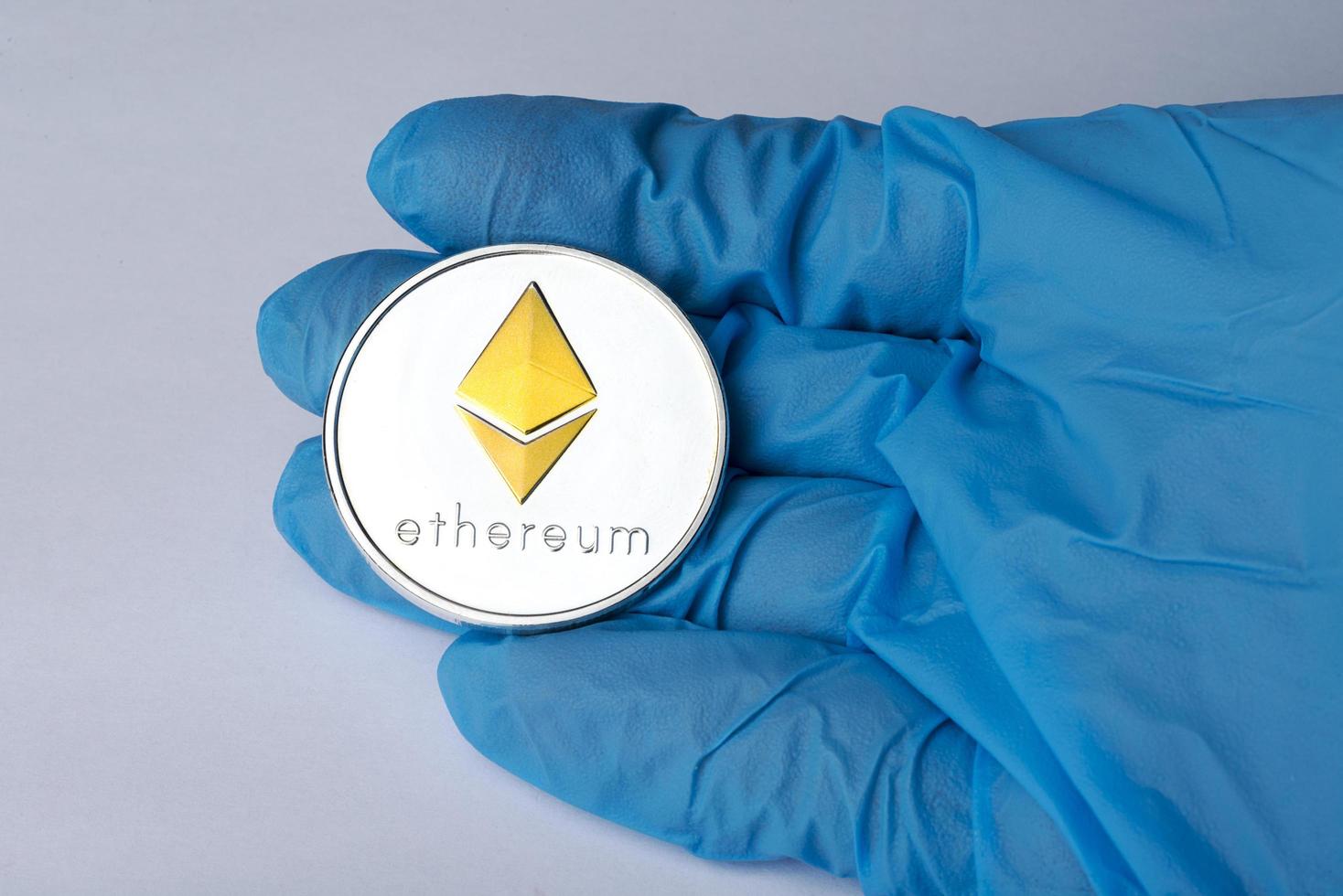 Hand in blue glove holding a silver Ethereum. photo