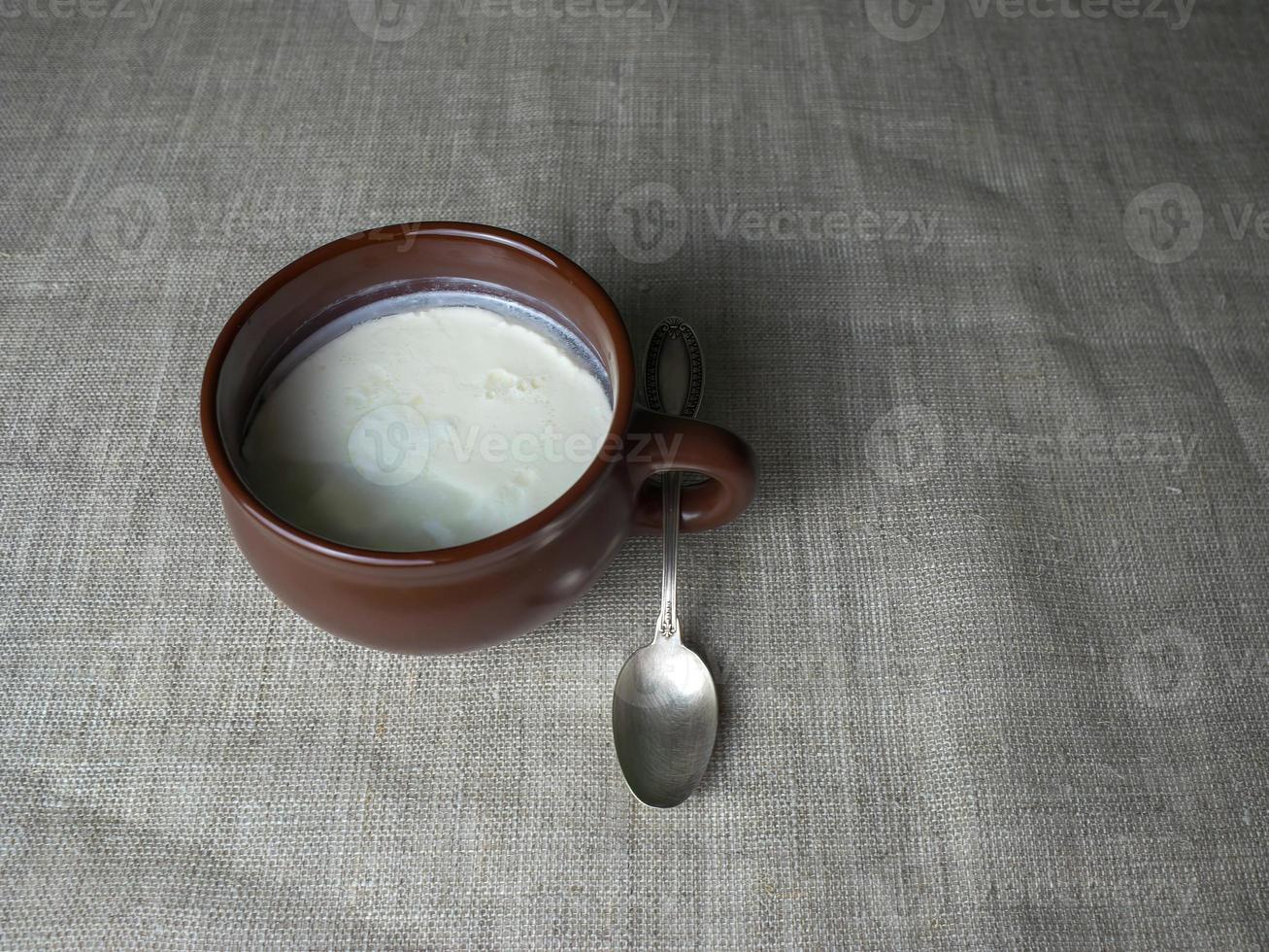 Plain white homemade yogurt in a brown clay cup, next to it is a silver spoon on a gray linen tablecloth. Minimalistic food background photo
