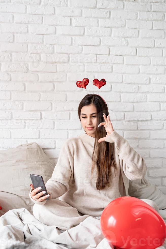 joven mujer divertida sentada en la cama celebrando el día de san valentín chateando usando el teléfono móvil foto