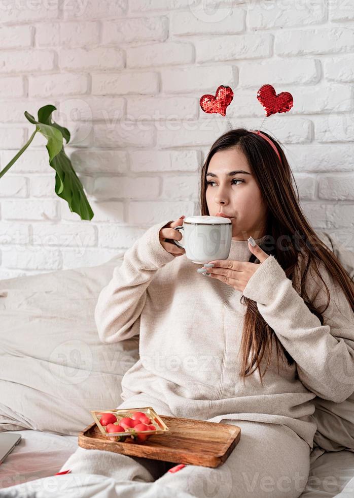 día de san valentín, día de la mujer. joven morena caucásica sentada en la cama celebrando el día de san valentín trabajando en una laptop en línea foto