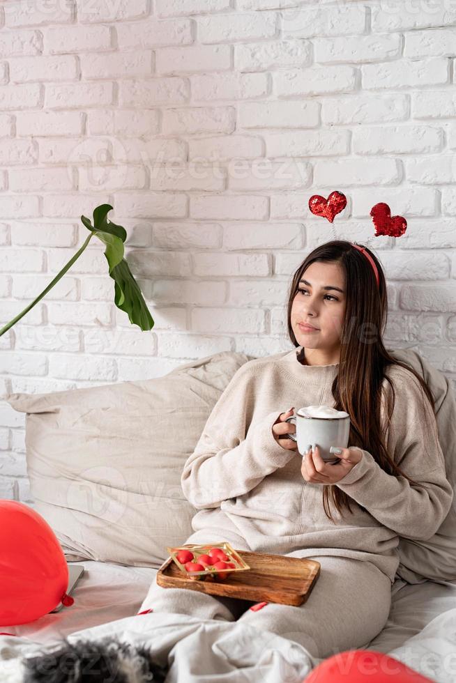 día de san valentín, día de la mujer. joven morena caucásica sentada en la cama celebrando el día de san valentín trabajando en una laptop en línea foto