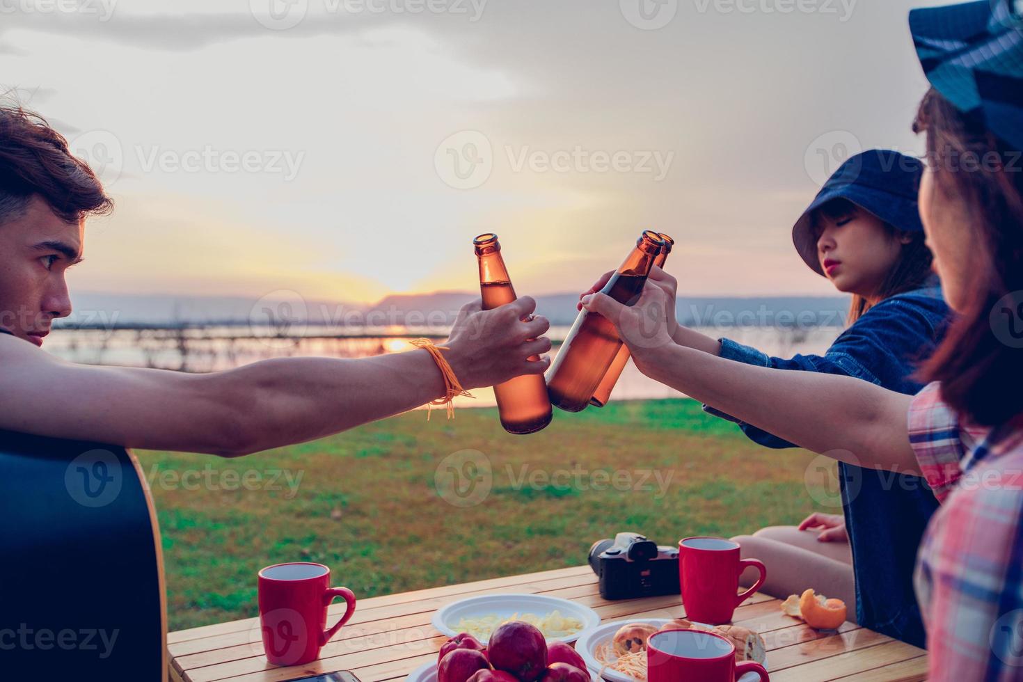A group of Asian friends tourist drinking alcoholic beer and playing guitar together with happiness in Summer while having camping near lake at sunset photo