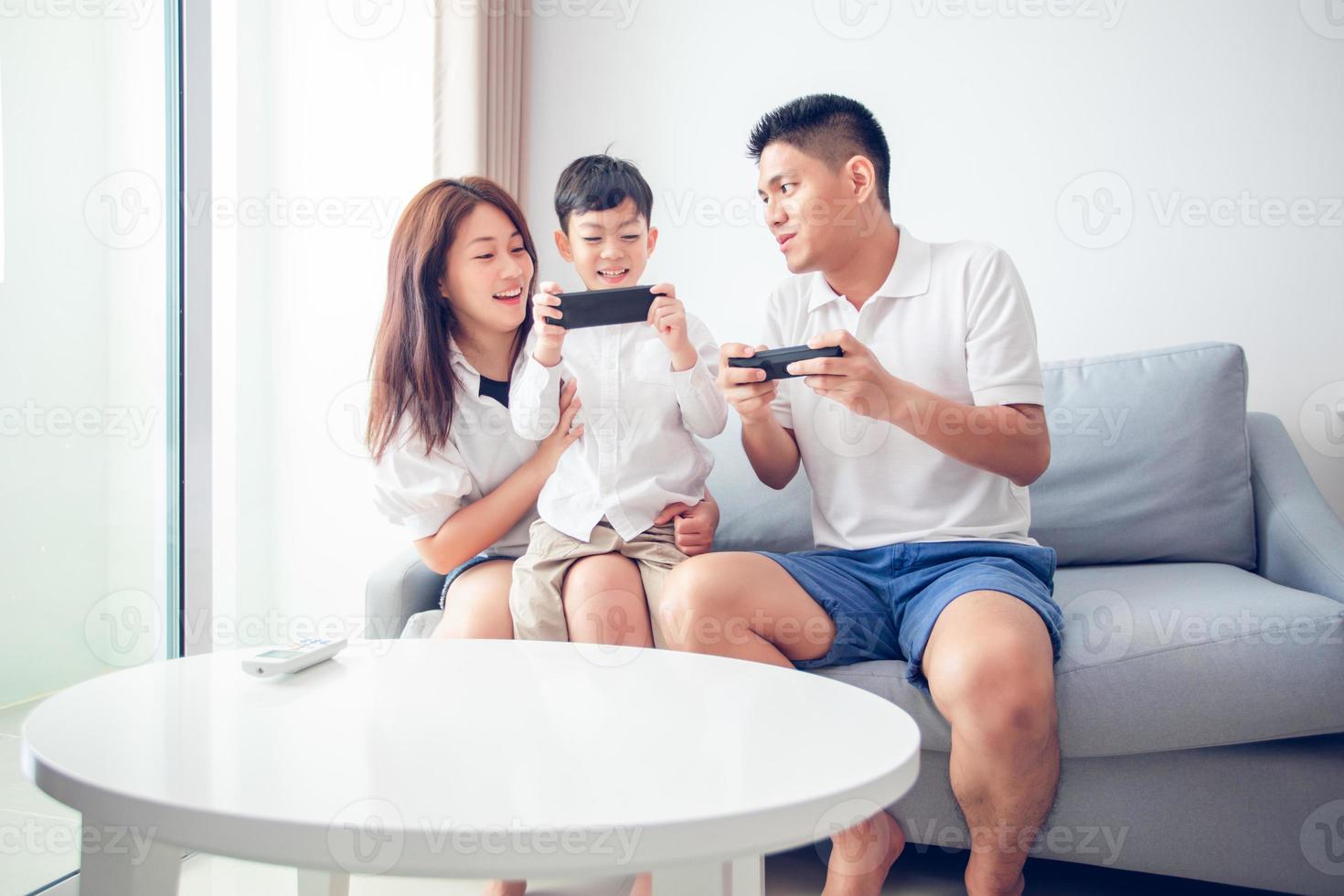 familia asiática divirtiéndose jugando juegos de consola de computadora juntos, padre e hijo tienen los controladores del teléfono y la madre anima a los jugadores. foto