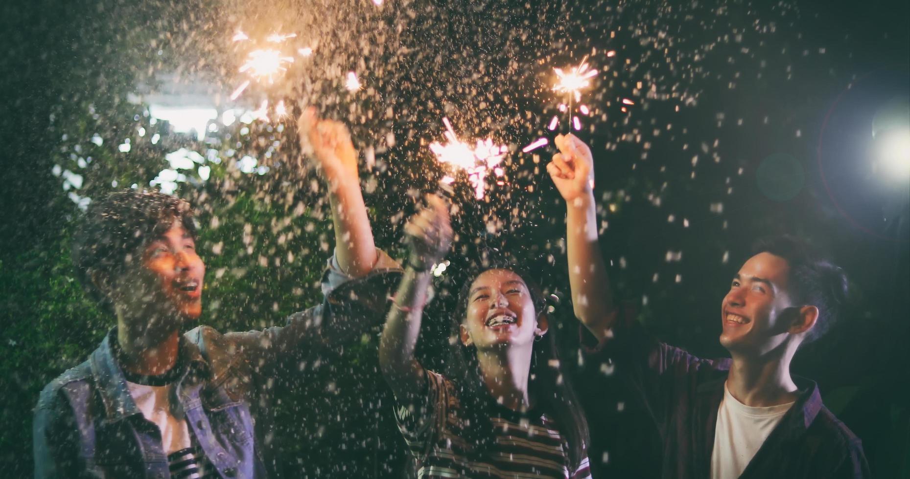 Grupo asiático de amigos con barbacoa en el jardín al aire libre riendo con bebidas alcohólicas de cerveza y mostrando un grupo de amigos divirtiéndose con bengalas en la noche, enfoque suave foto