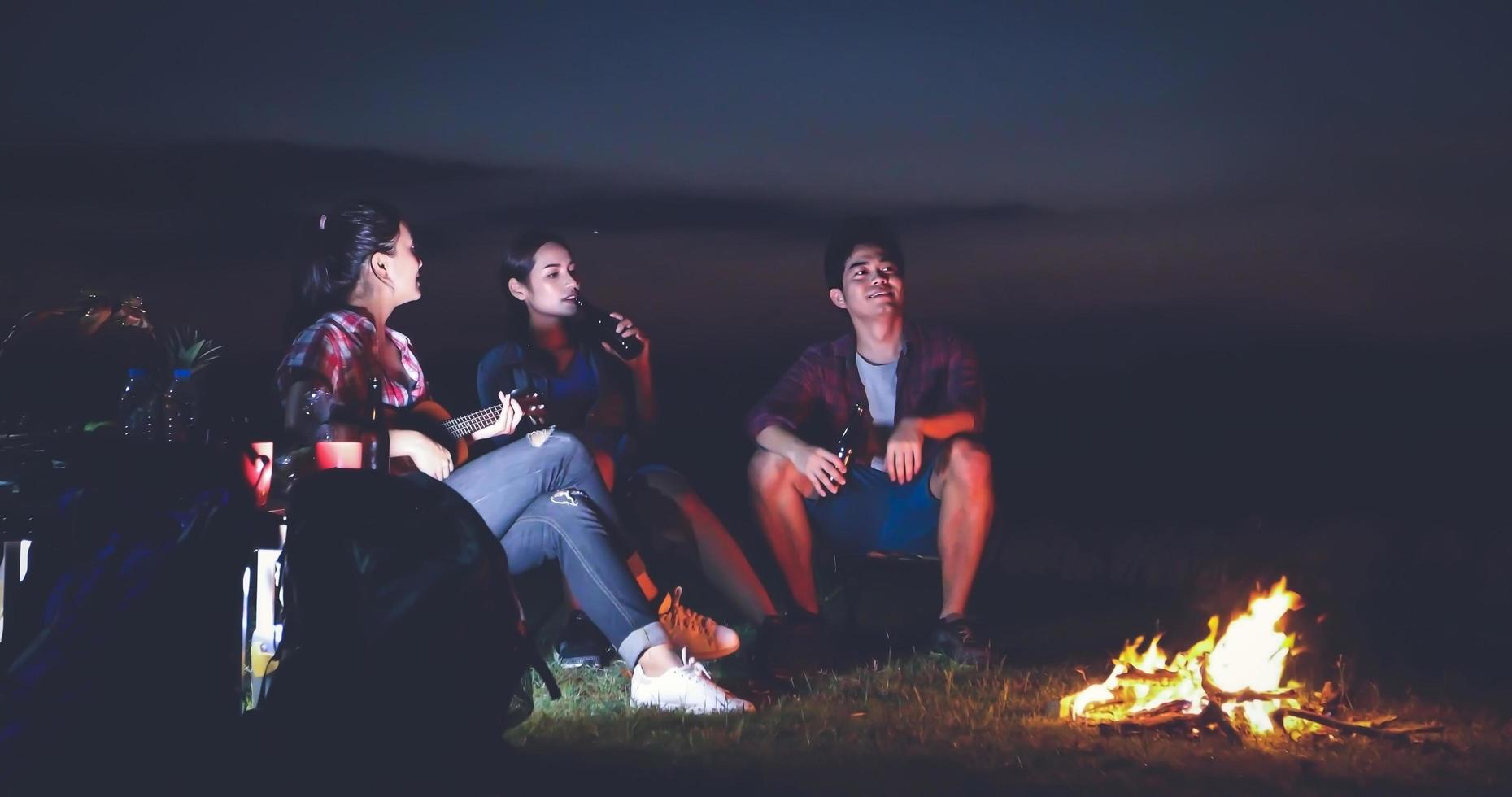 A group of Asian friends tourist drinking and playing guitar together with happiness in Summer while having camping near lake at sunset photo