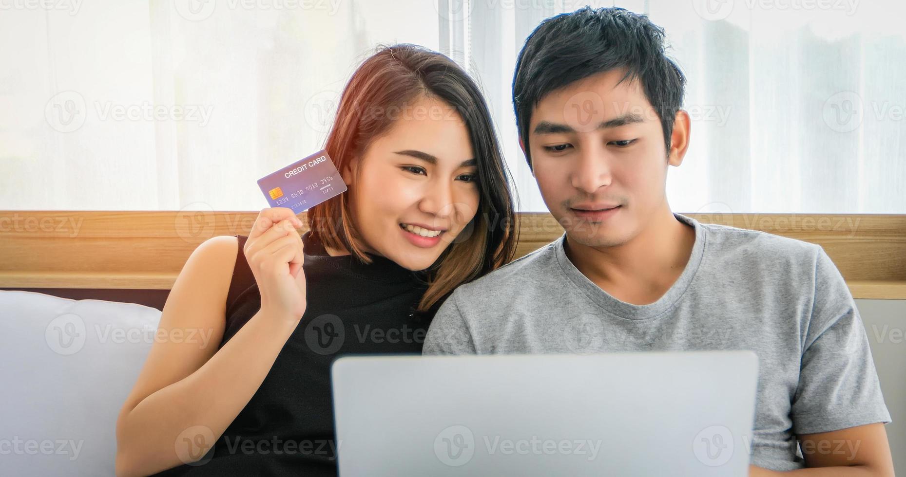 pareja asiática comprando en línea y pagando con tarjeta de crédito en una computadora portátil, pareja feliz en casa navegando por la red en la cama foto