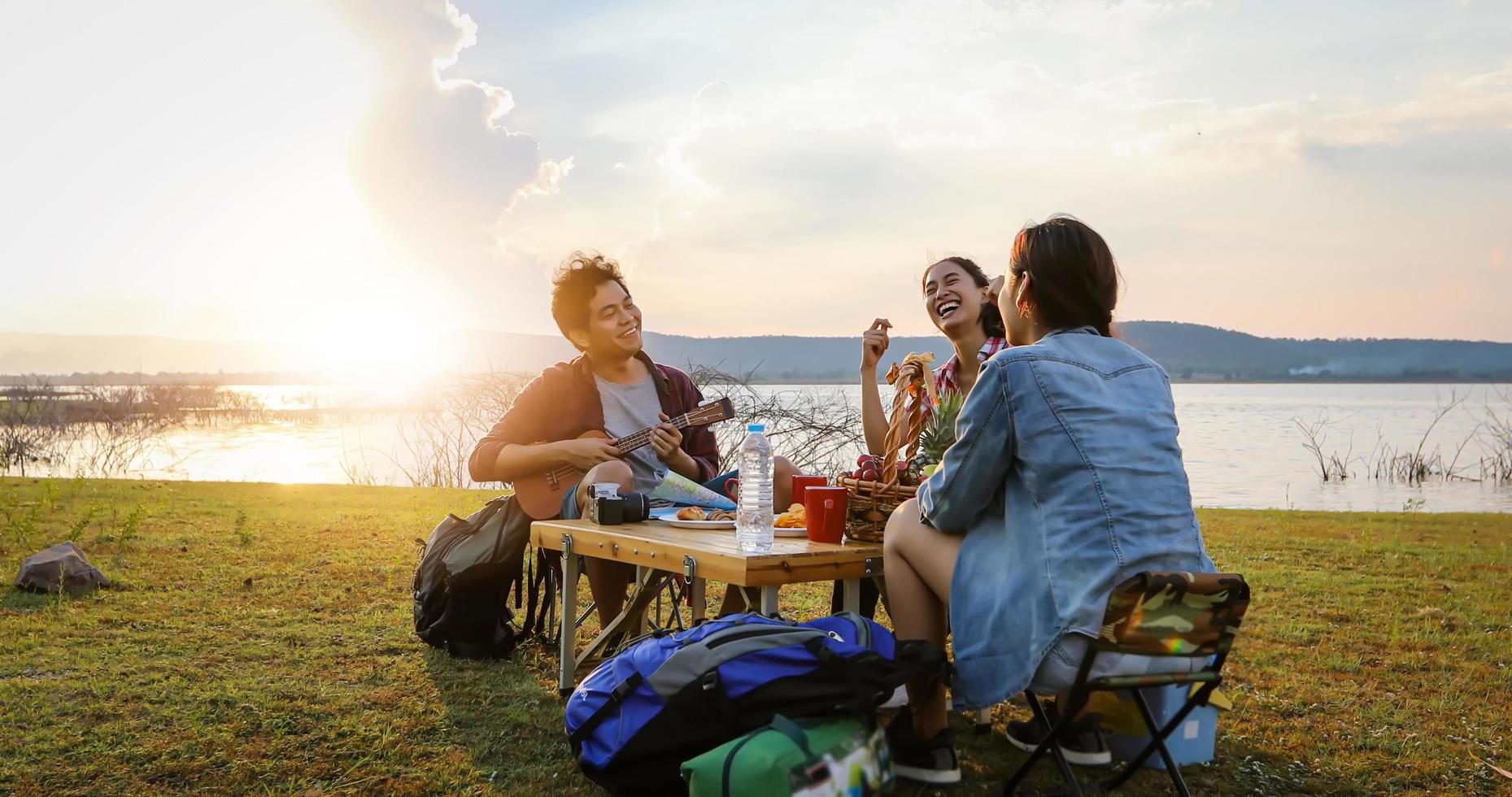 A group of Asian friends tourist drinking and playing guitar together with happiness in Summer while having camping near lake at sunset photo