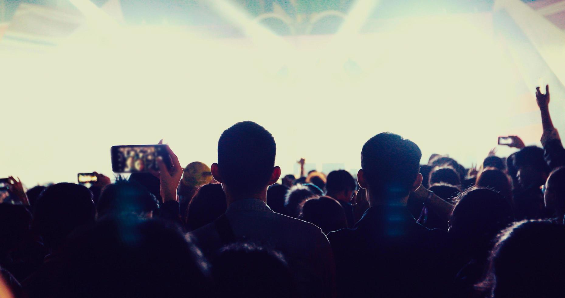 Siluetas de multitud de conciertos en la vista trasera de la multitud del festival levantando sus manos sobre las luces del escenario brillante foto