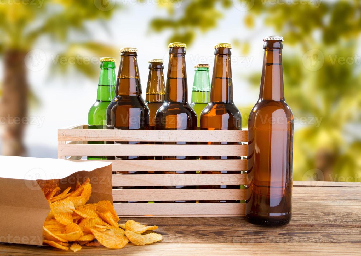 many beer bottles and pack of chips on wooden table on blurred tropical background photo