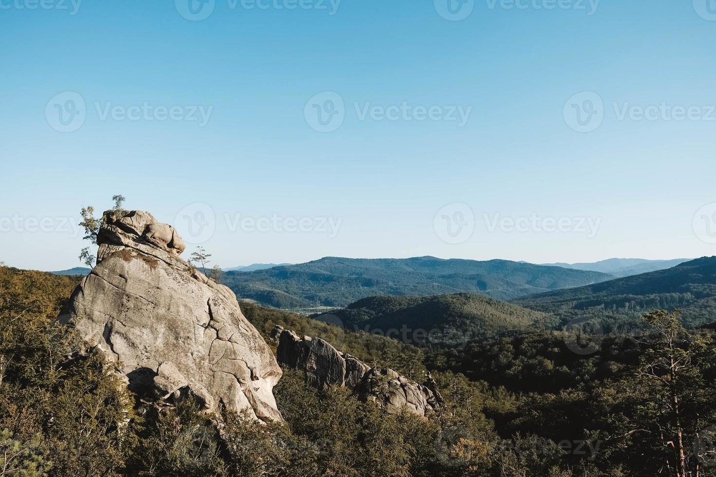 Landscape with high stone rock in a green forest under a blue sky on a background of mountains. Copy, empty space for text photo