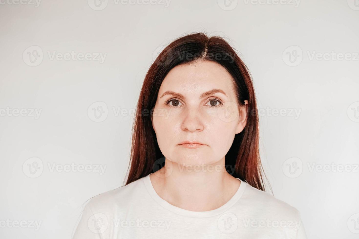 Portrait of a serious woman dressed in casual white t shirt, has minimal makeup, poses indoor on a white background. Copy, empty space for text photo