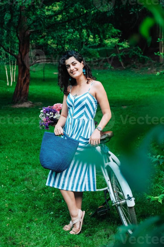 Woman dressed in a in a striped dress is standing next to a bicycle with a basket of flowers in park photo