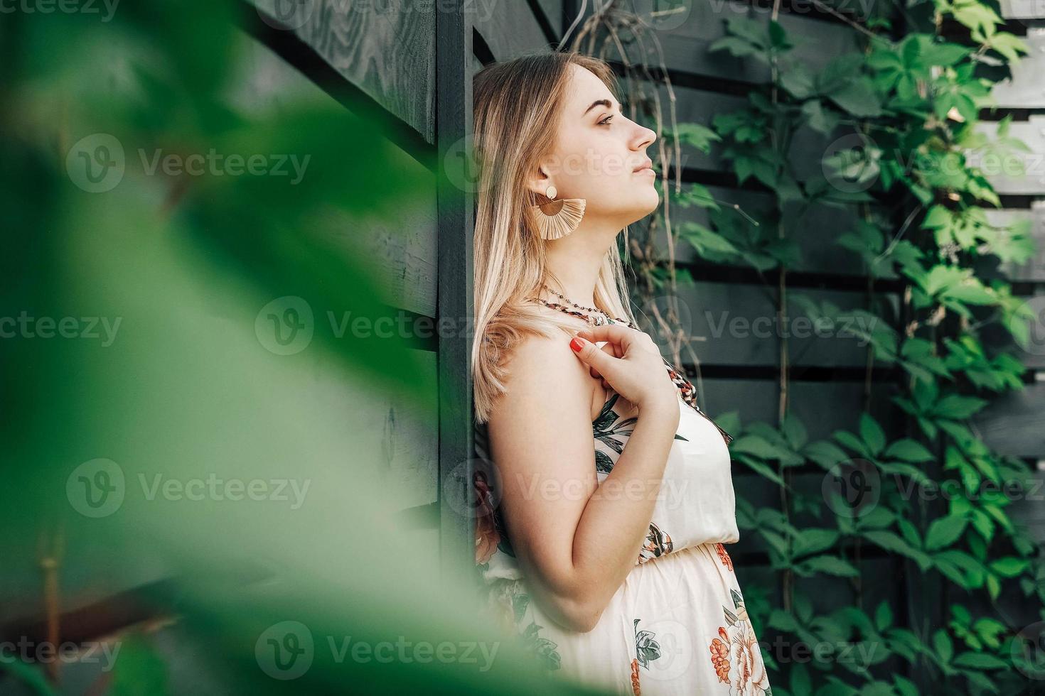 Portrait of a beautiful blonde woman dressed in flower dress in a green leaves trees on a wooden wall background. Copy, empty space for text photo