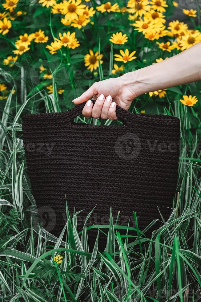 Woman's hand holding a brown knitted bag on a background of grass and flowers. Copy, empty space for text photo