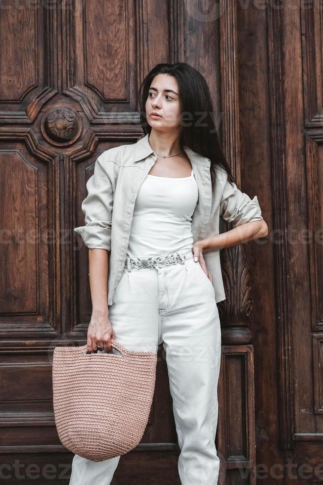 Woman wearing in white clothes, holding knitted bag on the background of old wooden doors. Copy, empty space for text photo