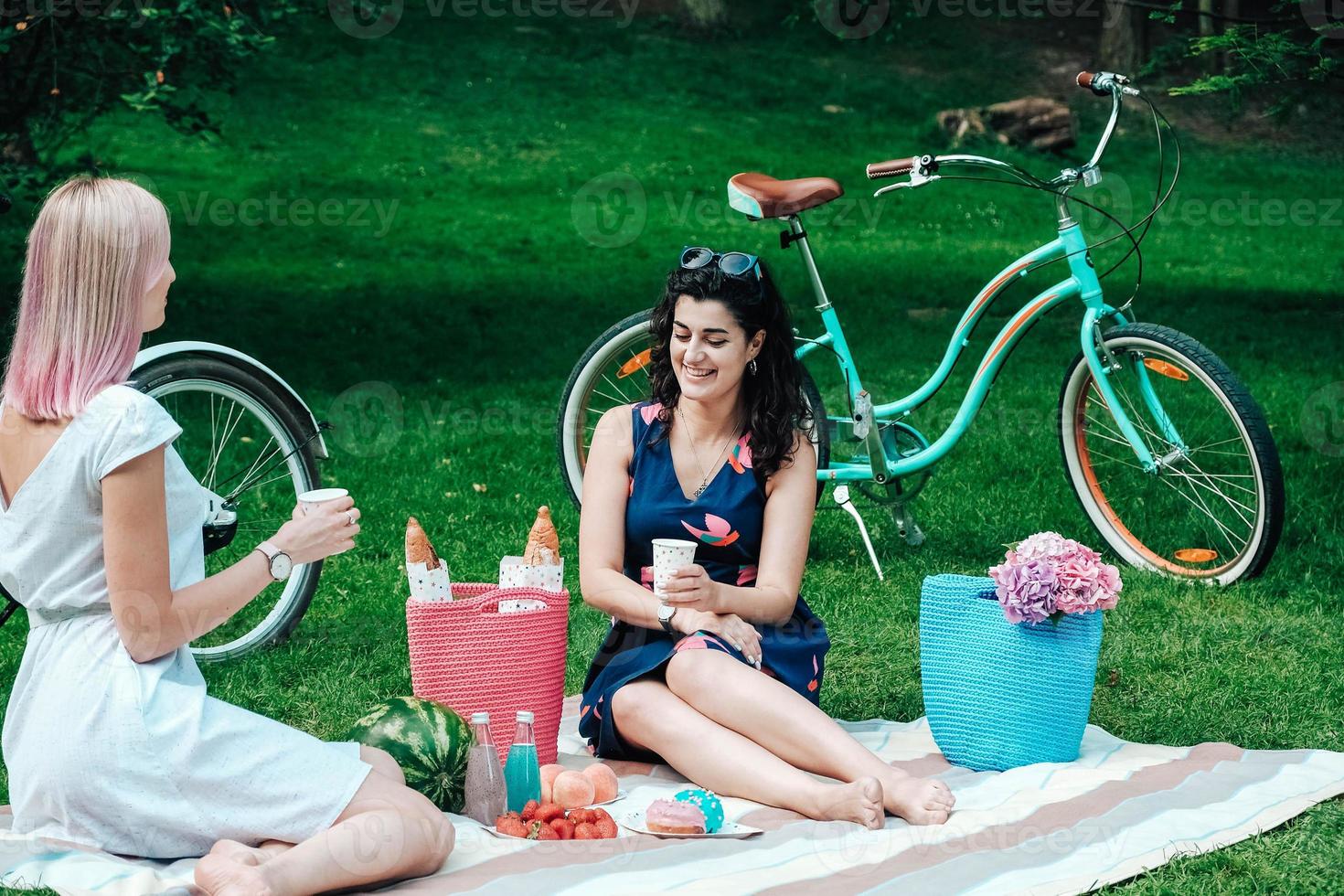 Two women in trendy summer sundress sitting on a plaid in park on background of a bicycle photo