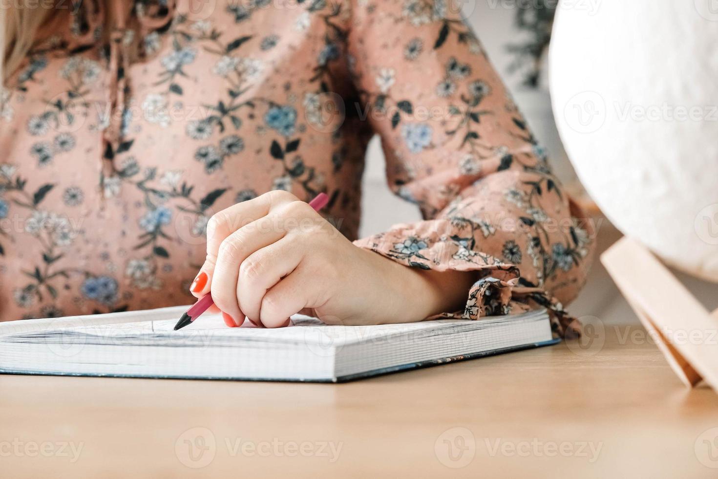 Woman hands with pencil, she is is writing in her notebook sitting at the table with book shelf background. Copy, empty space for text photo