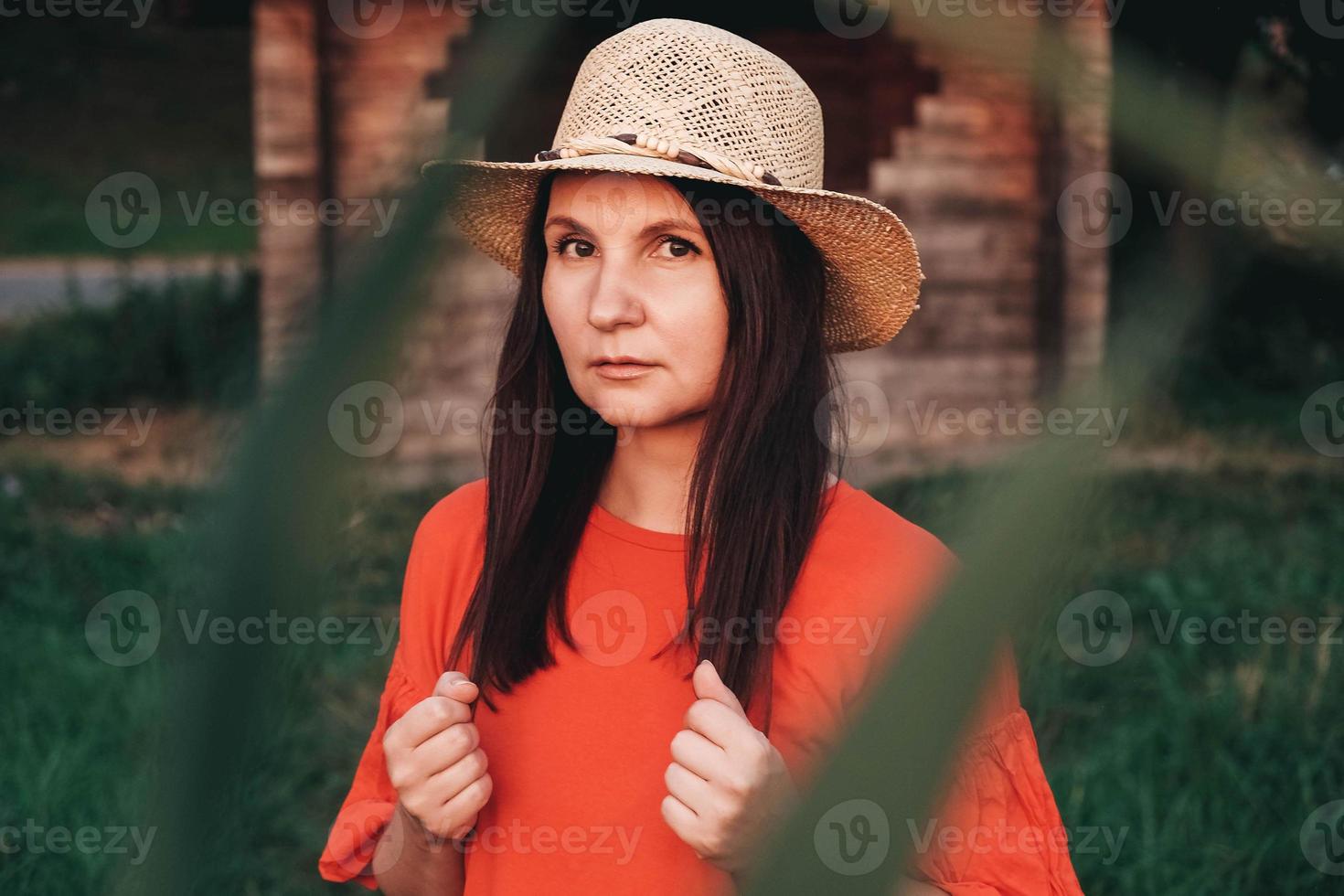 Portrait of a beautiful woman in a straw hat dressed in red clothes stands on the background of a wooden house photo
