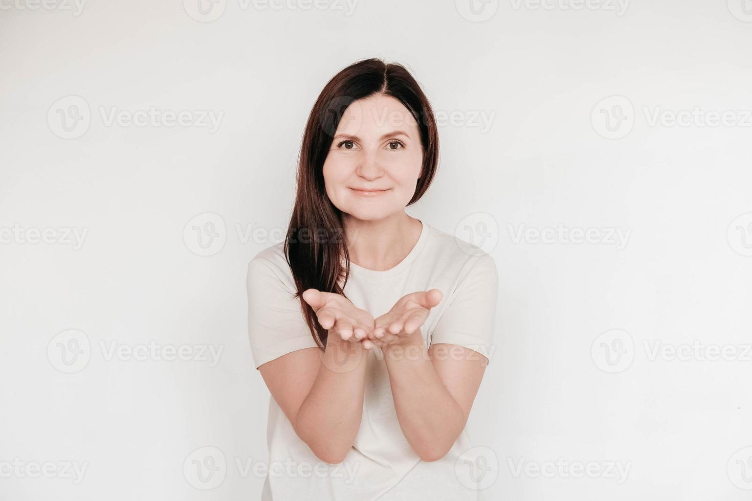 Beautiful brunette woman keeps hands on cheek, dressed in casual white t shirt, has minimal makeup, poses indoor on a white background photo