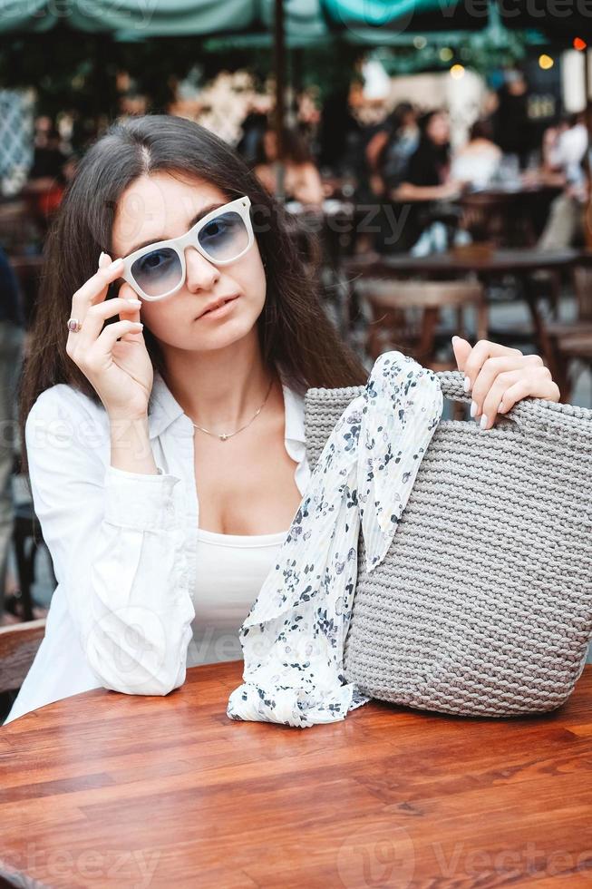 mujer con gafas de sol y ropa blanca, sosteniendo una bolsa de punto sentada en la mesa de un café de la calle. copiar, espacio vacío para texto foto