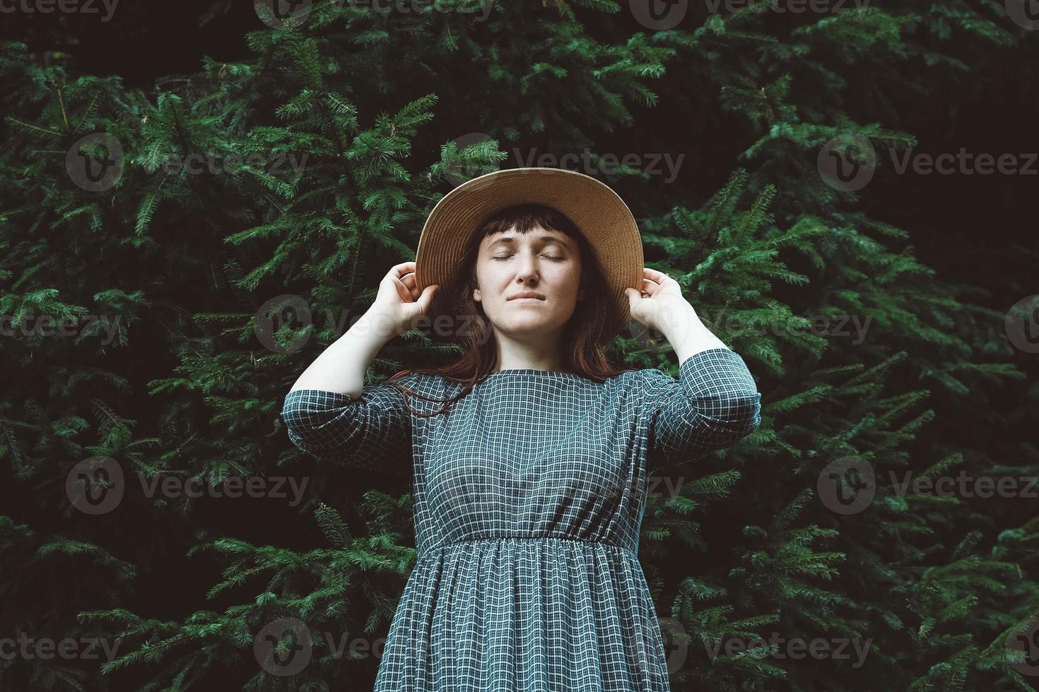 mujer con sombrero de paja y vestido con los ojos cerrados sobre un fondo de bosque verde y árboles foto