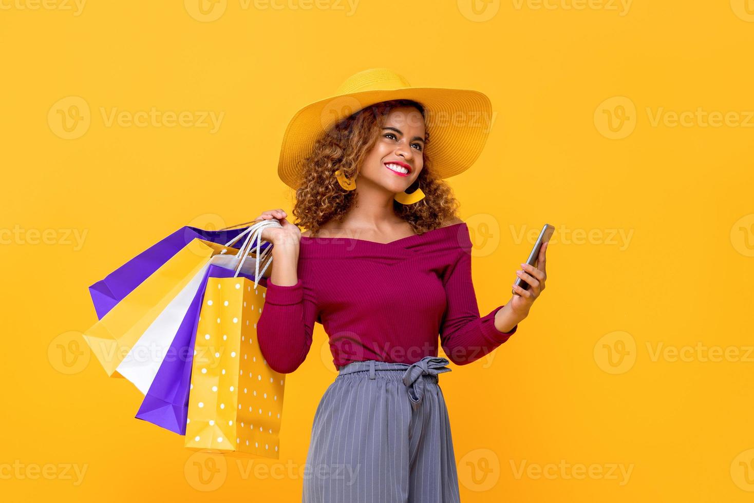 Fashionable smiling mixed race woman with colorful shopping bags and mobile phone isolated on yellow background for summer sale conept photo