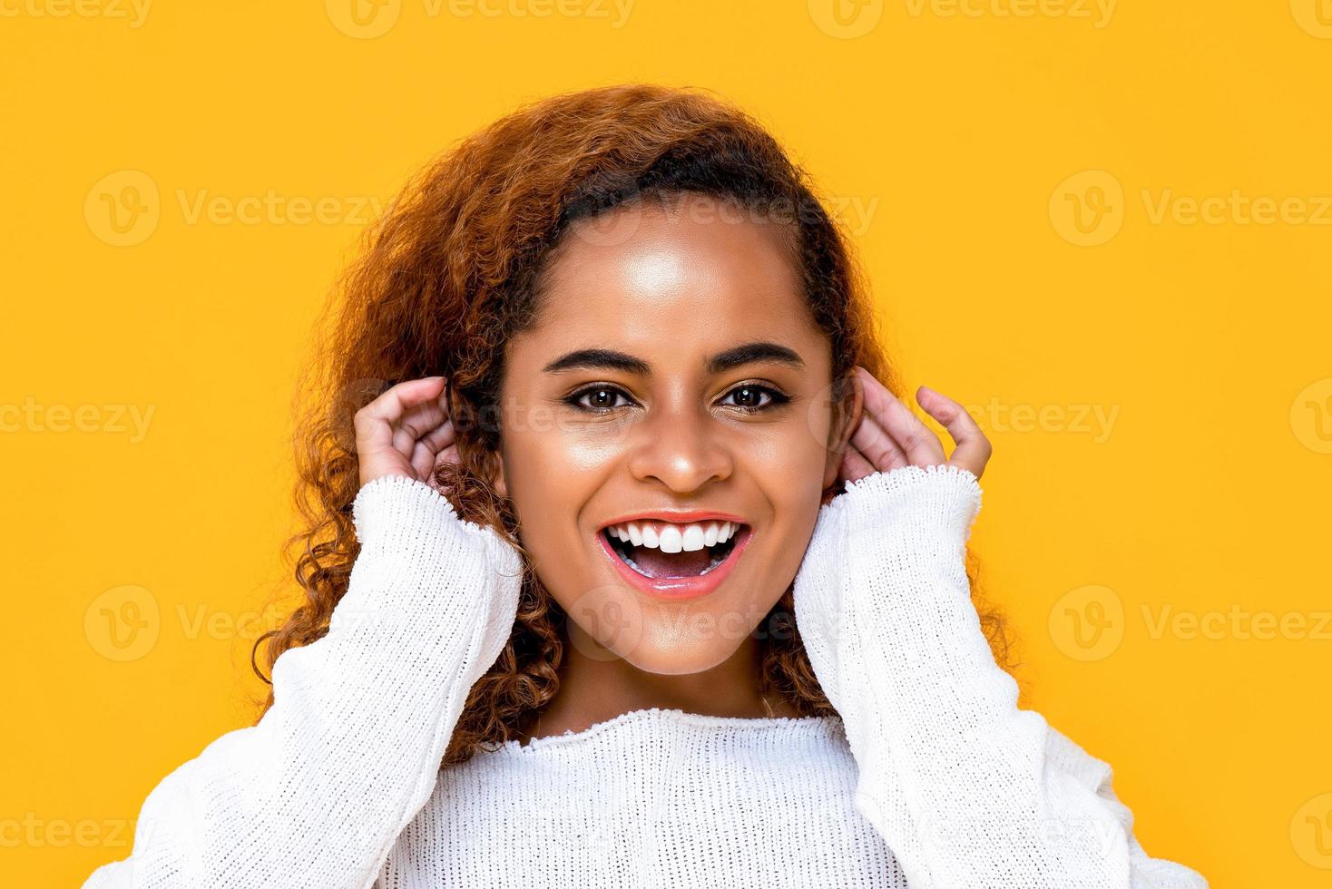 primer plano retrato de una alegre joven afroamericana sonriendo mientras toca sus oídos con ambas manos en un estudio aislado de fondo amarillo foto