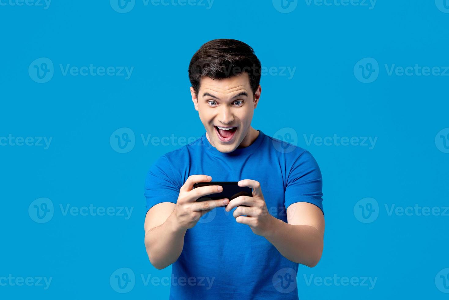 Excited handsome young male gamer playing online game on smartphone isolated on blue background photo