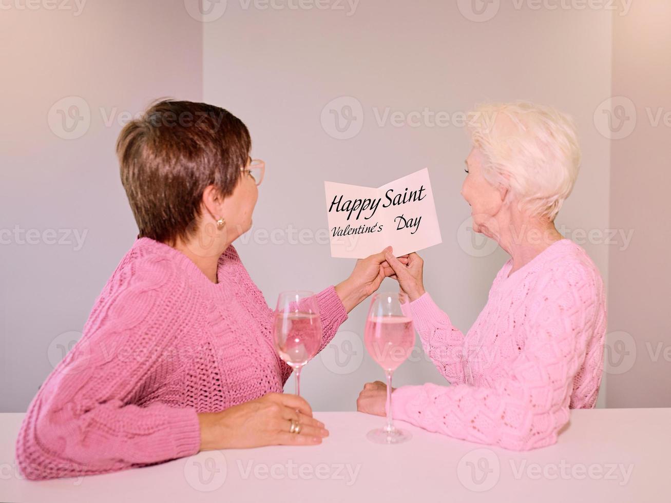 Two mature women drinking wine and giving a post card. Friendship, holidays, love, same-sex marriage concept photo