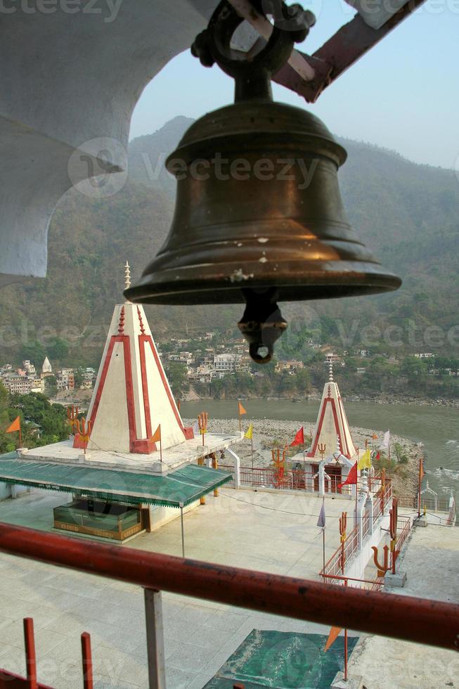 Temples on the banks of river Ganga with brass bell in foreground, Rishikesh, Uttarakhand, India, Asia photo