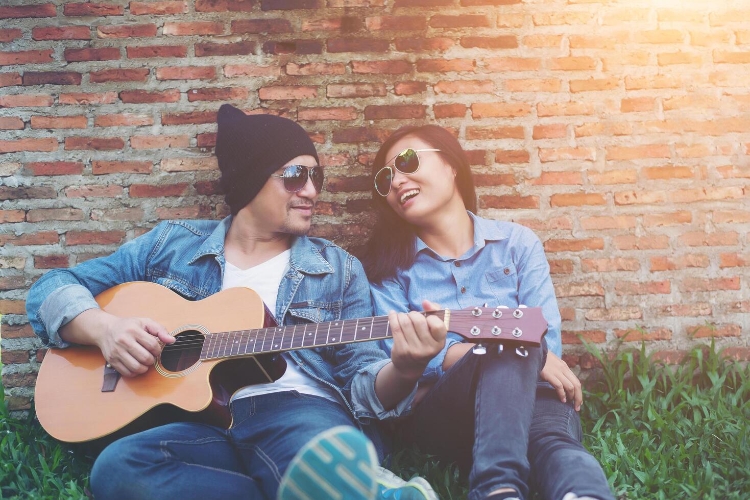 Hipster man playing guitar for his girlfriend outdoor against brick wall, enjoying together. photo