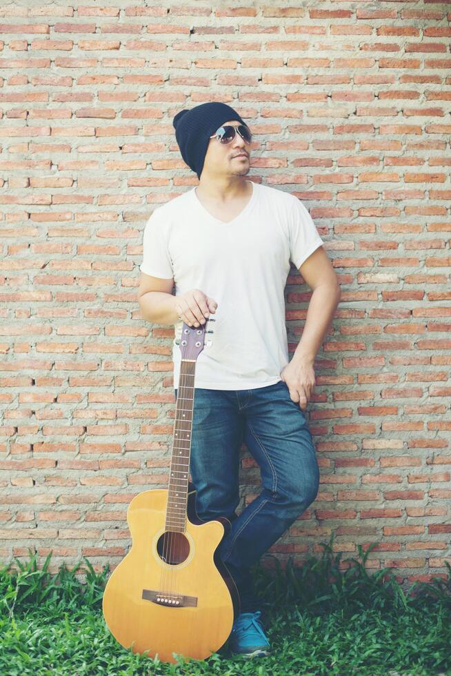 Handsome guy standing holding guitar against the brick wall posing look away. Relaxing holiday. photo