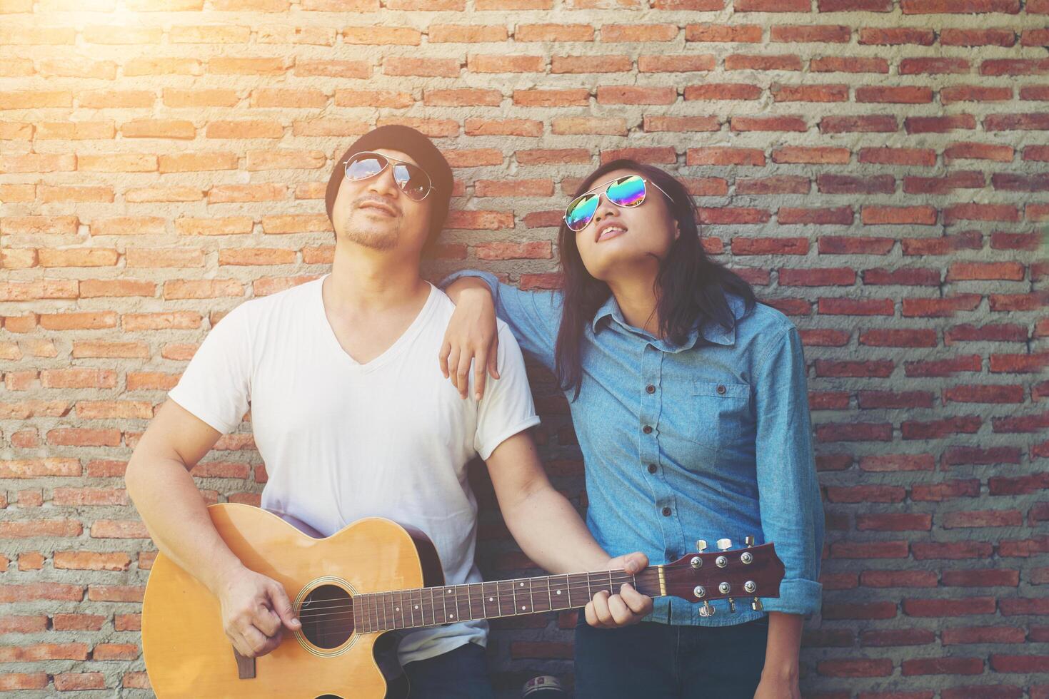 Hipster couple is hugging, looking away and smiling while standing outdoor playing guitar against brick wall, Dating spent great time together. photo