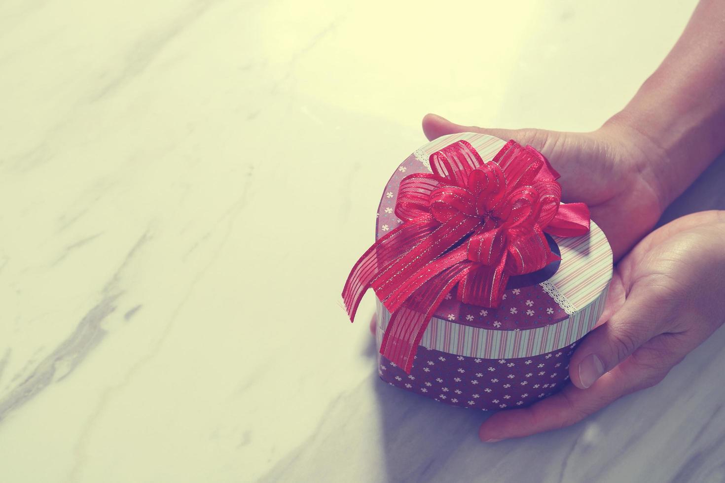 entrega de regalos, mano de hombre sosteniendo una caja de regalo en forma de corazón en un gesto de dar sobre fondo de mesa de mármol gris blanco foto